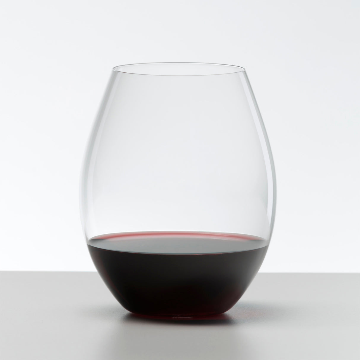 A Riedel O Range Stemless Old World Syrah Glass from Riedel, partially filled with red wine, rests on a light gray surface against a plain white background. This stylish set of two crystal glasses (0414/41) is both elegant and dishwasher safe.