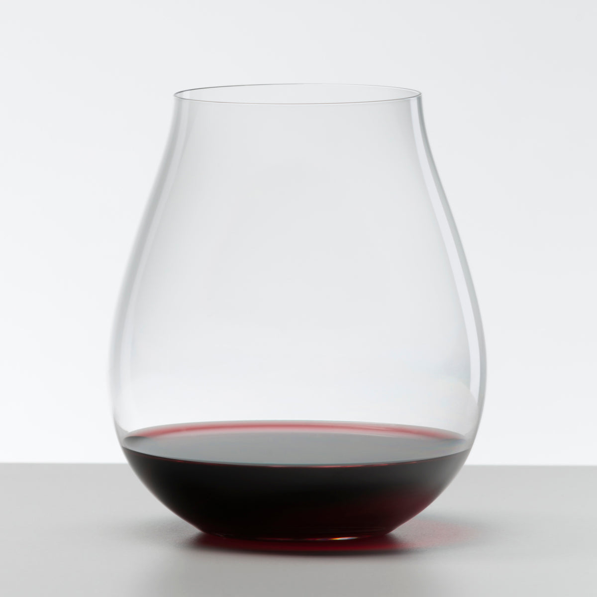 A clear, tulip-shaped stemless glass from the Riedel O Range Stemless New World Pinot Noir set (0414/67) by Riedel holds a small amount of red wine on a white surface with a plain background.