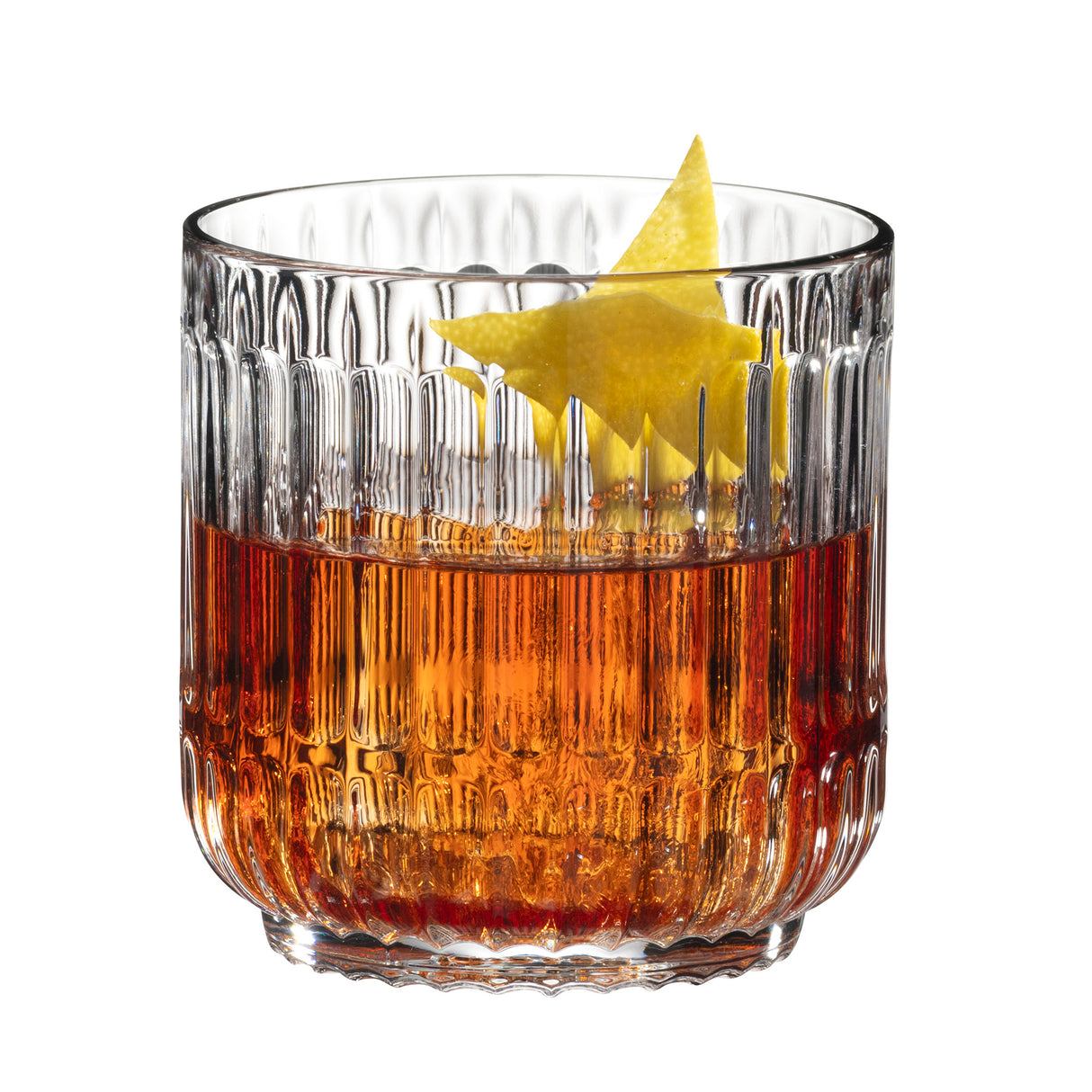 A Riedel Restaurant Bar Rituals Rocks Tumbler (283ml) by Riedel, crafted from lead-free crystal with vertical grooves, is filled with an amber-colored drink and garnished with a twist of lemon peel. The glass sits on a white background.
