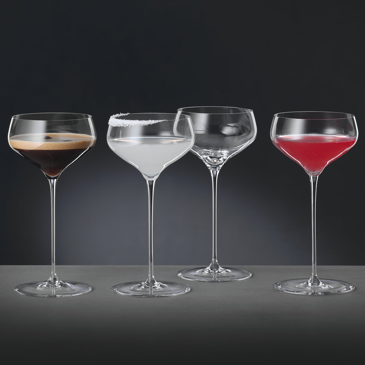 Four Spiegelau Hi-Lite Coupette / Cocktail Saucer glasses from Spiegelau are lined up in a row—featuring both a crystal cocktail glass and an elegant coupe glass—each holding a different drink: one with a dark foamy cocktail, another with a clear beverage topped by a salt rim, one empty glass, and one filled with a red cocktail. The arrangement rests on a grey surface set against a dark background.