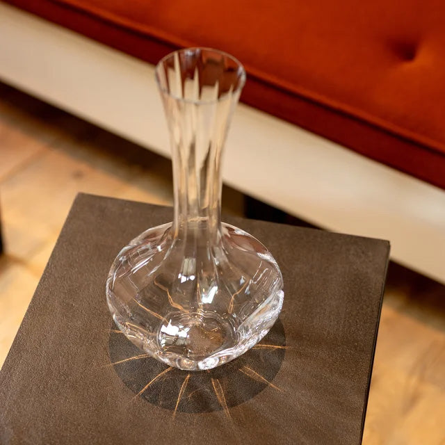 A Riedel Manufaktur Exquisit Wine Decanter 1.2L by Riedel sits on a brown tabletop, its clear, faceted crystal design casting a shadow. In the background, part of an orange cushioned seat with white trim is visible.