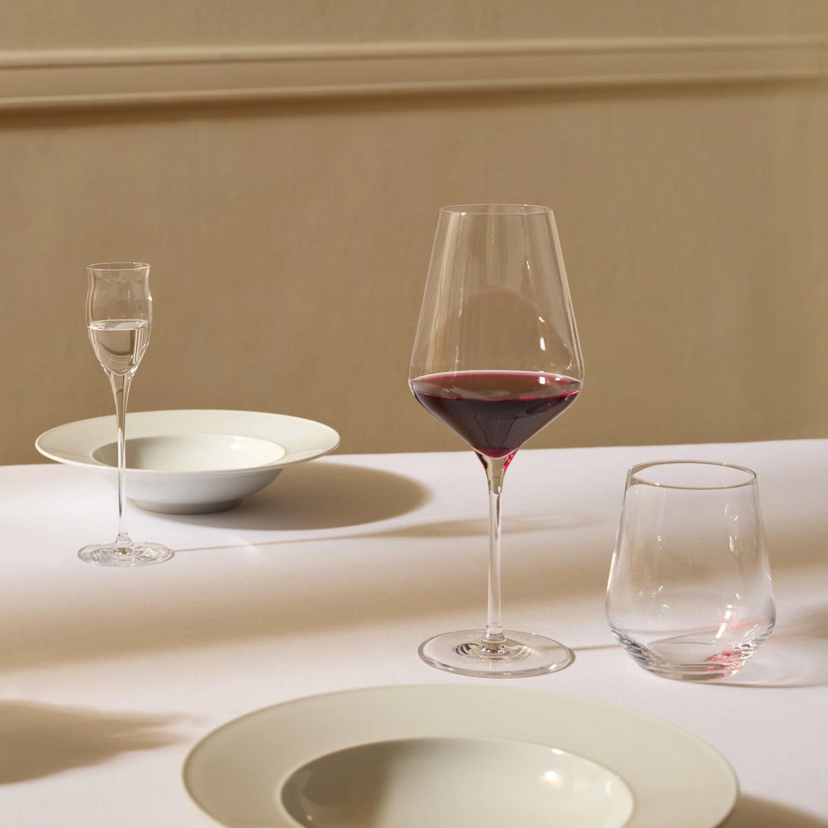 A table setting featuring a tall, angular Stolzle Quatrophil Red Wine Glass filled with wine, accompanied by a small stemmed glass, a short tumbler, and two wide-rimmed bowls.