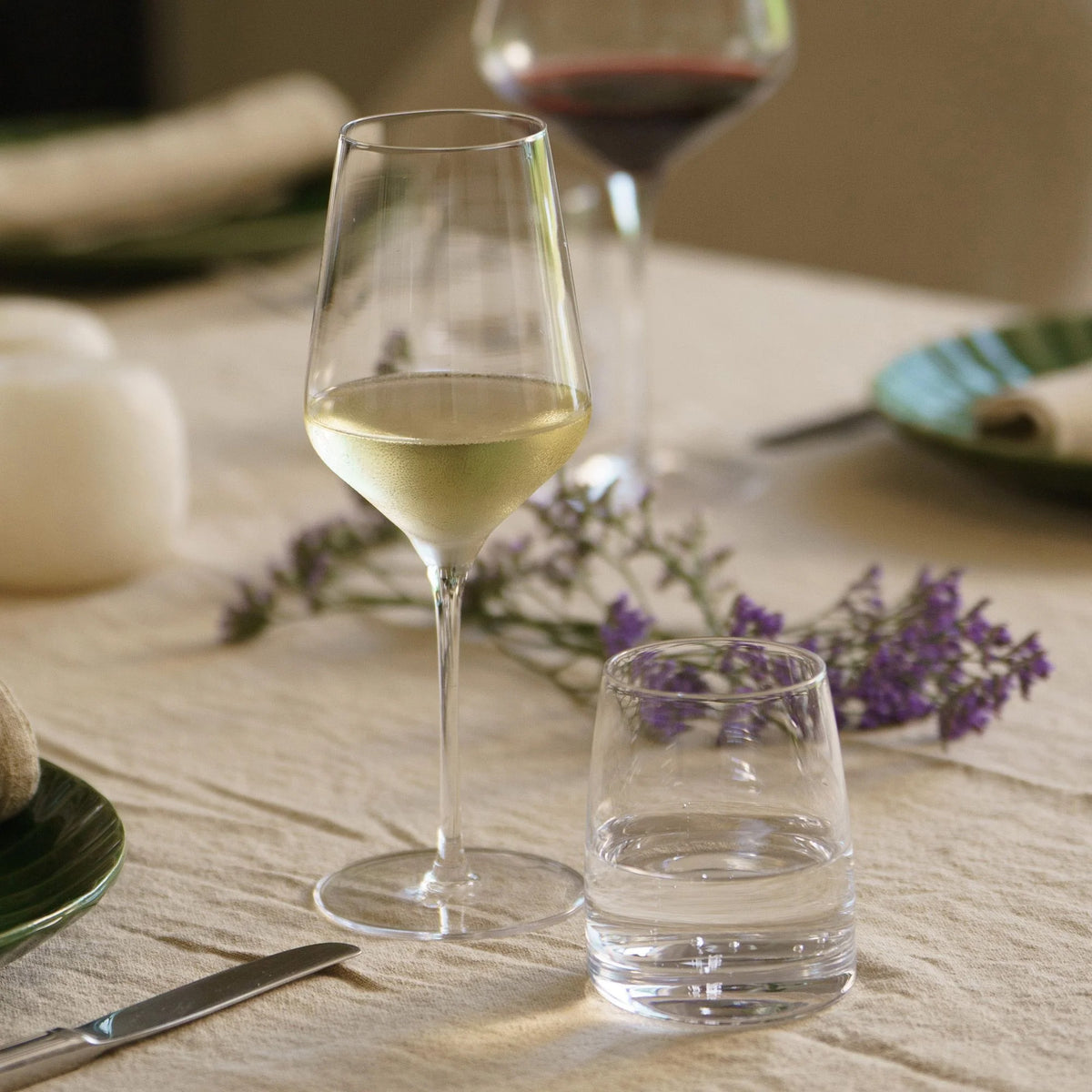 A table set with Stolzle STARlight White Wine Glass - Set of 6, water glasses, plates, cutlery, and purple flowers on a beige tablecloth exudes elegance for serving white wine.