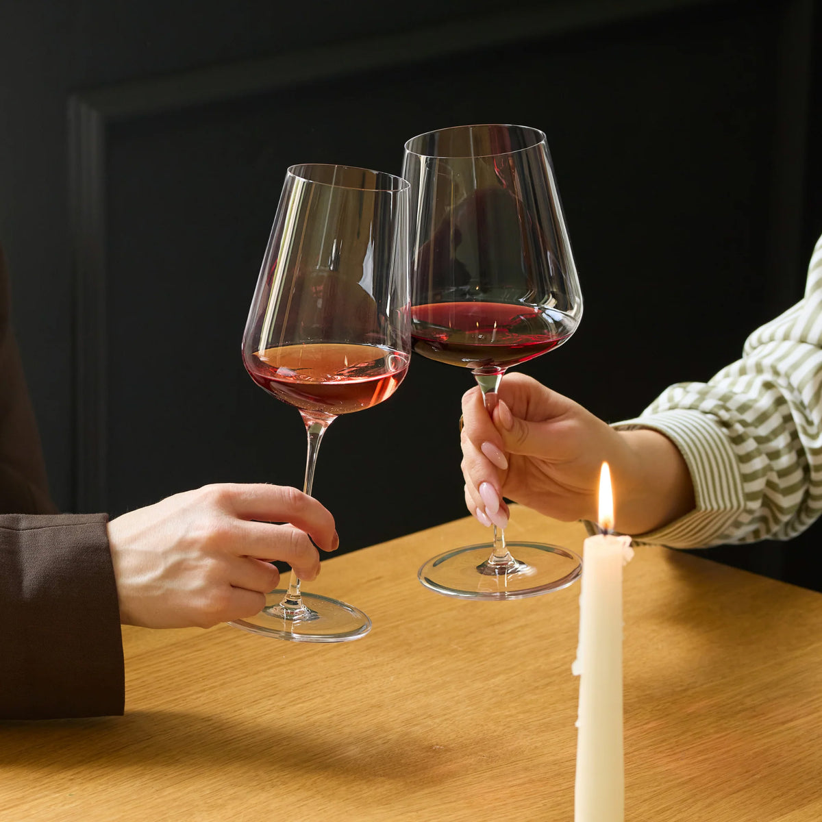 Two people clink Stolzle STARlight Universal Red & White Wine Glass crystal wine glasses filled with red and white wine over a wooden table with a lit candle.