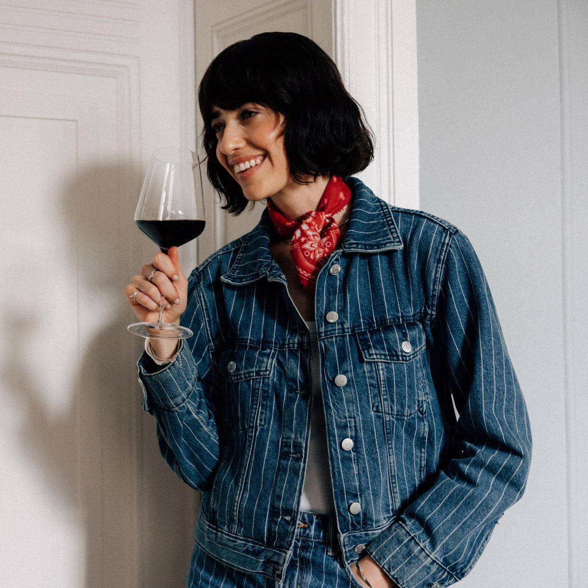 A smiling woman in a denim jacket and red bandana holds a Stölzle STARlight Bordeaux Red Wine Glass filled with red wine.