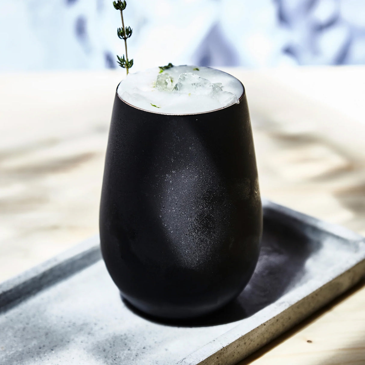 The Stölzle Elements Black & Bronze Water/Mixer Tumbler Glass 465ml, shown filled with a frothy iced drink and a greenery garnish on a tray, features sleek black design. Sold as a set of 6.