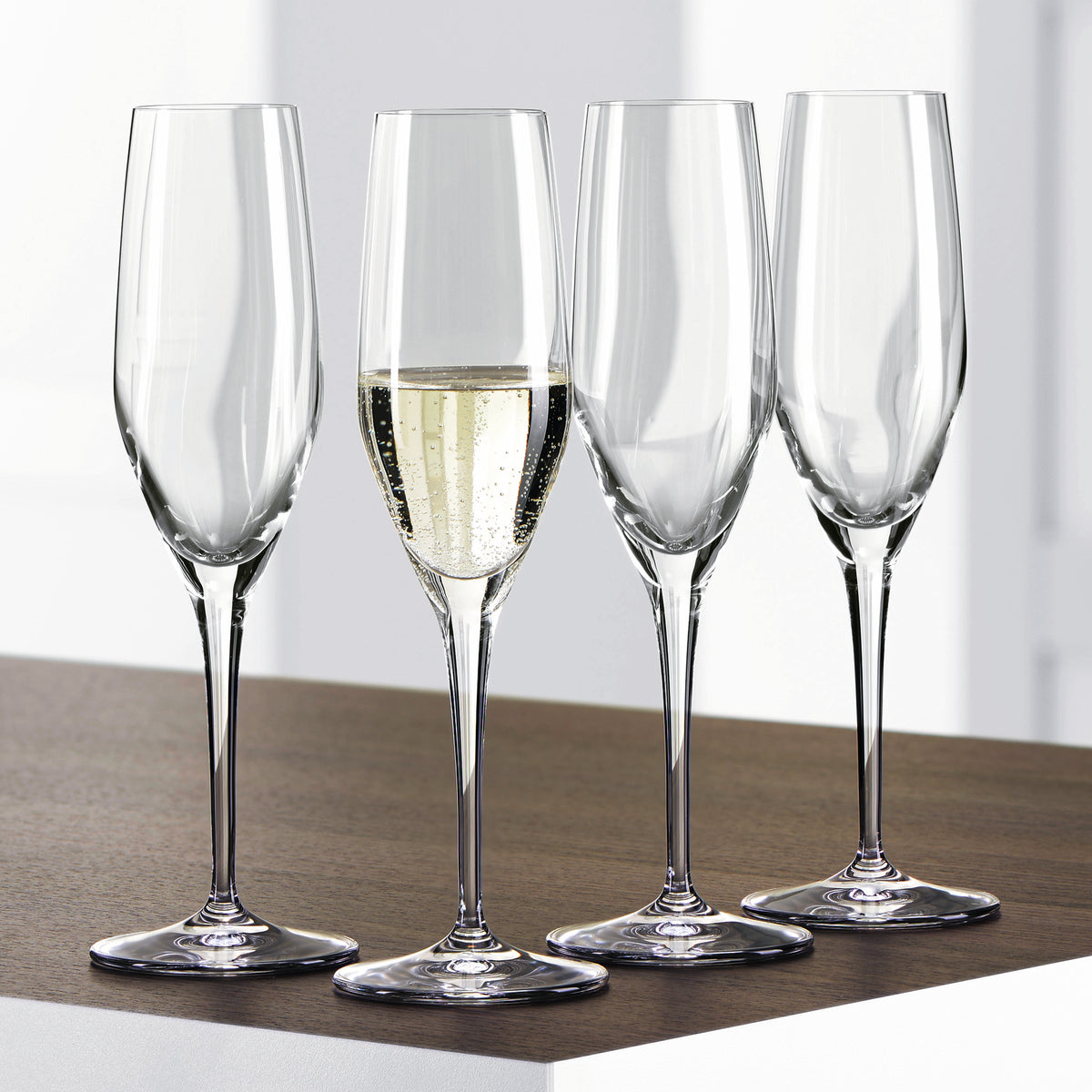 Twelve Spiegelau Restaurant Authentis Champagne Glasses (220ml) from Spiegelau are elegantly arranged on a wooden surface; three of the exquisite crystal glasses are empty and one is filled with sparkling champagne. The softly lit, out-of-focus background enhances the sophisticated presentation.