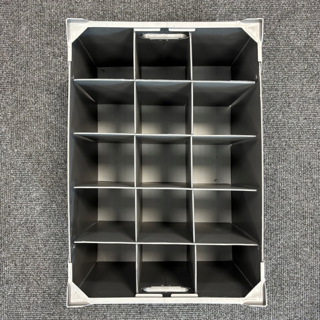 Top-down view of an empty Wineware USED - GREAT CONDITION Wine Glass Storage Box, featuring fifteen rectangular compartments arranged in a 3 by 5 grid, placed on a textured gray carpet. The dividers keep glassware separated, making this stackable 240mm high organizer ideal for safely storing and organizing delicate wine glasses.