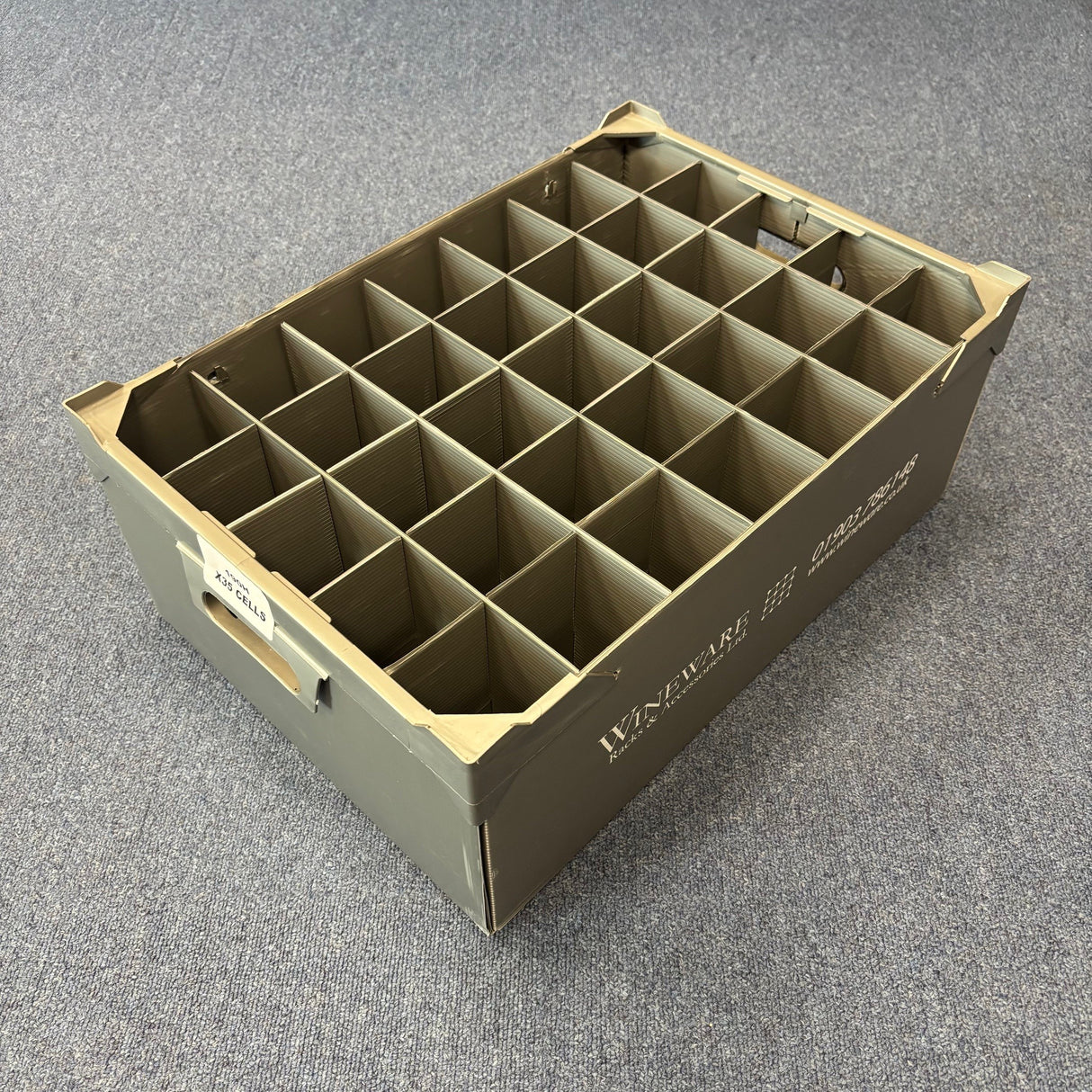 The Wineware USED - GREAT CONDITION Wine Glass Storage Box (190mm high, 35 cells) is a gray plastic organizer with handles and white side labels, ideal for safely storing delicate glassware. Seen here on a carpeted floor.