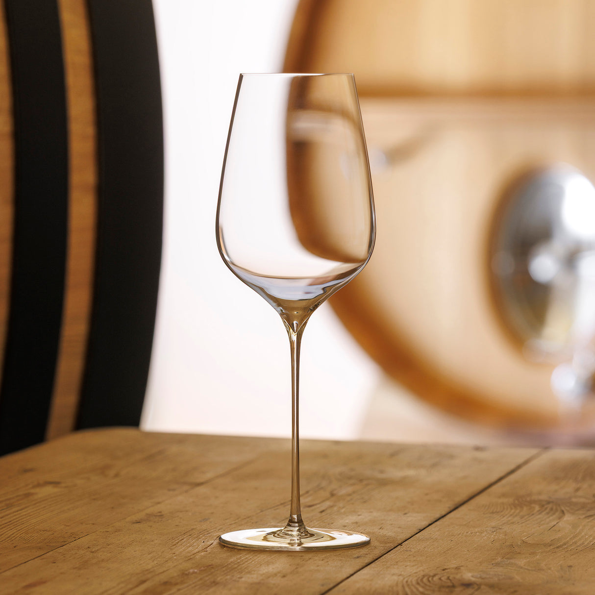 A pair of Sydonios Terroir Collection - Empreinte Red, White & Champagne Wine Glasses sit on a wooden table, showcasing elegant crystal design and oenological precision against softly blurred barrels in the background.