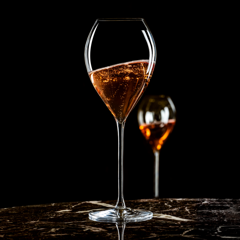 A Lehmann Glass Jamesse Grand Champagne / Sparkling Wine Glass (300ml) sparkles with rosé on a dark marble surface, bubbles rising. Another matching glass from the 6-piece set by Lehmann Glass is blurred in the background against a black backdrop.