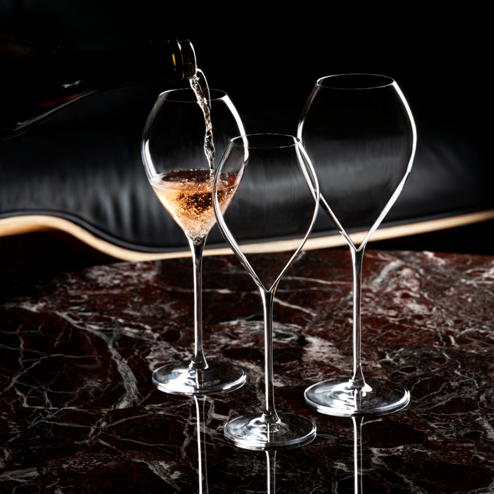 A bottle pours sparkling wine into a Lehmann Glass Jamesse Grand Champagne 300ml glass from the set of 6, with two empty crystal glasses nearby on a polished dark marble table against a blurred black background.