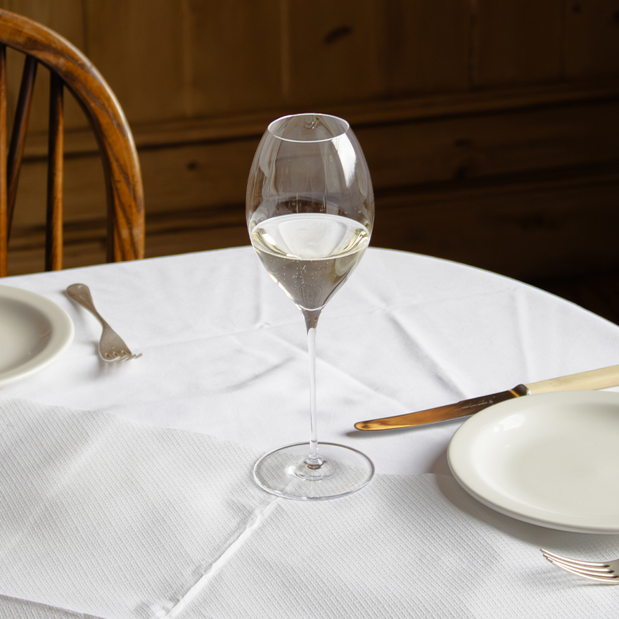 A Jancis Robinson x Richard Brendon The Occasional Collection Champagne/Sparkling Wine Glass - Set of 2 filled with white wine sits on a round table with a white tablecloth, two plates, cutlery, beside a wooden chair and paneled wall.