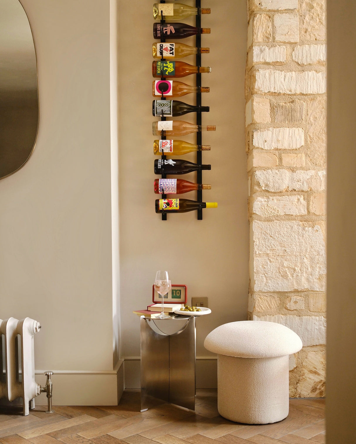 A modern interior features a VintageView Wall Mounted W Series 4 – 1220mm (12 to 36 Bottle Wine Rack) displayed on the wall, a small table with a wine glass and book, and a round cream stool positioned next to a textured stone wall.