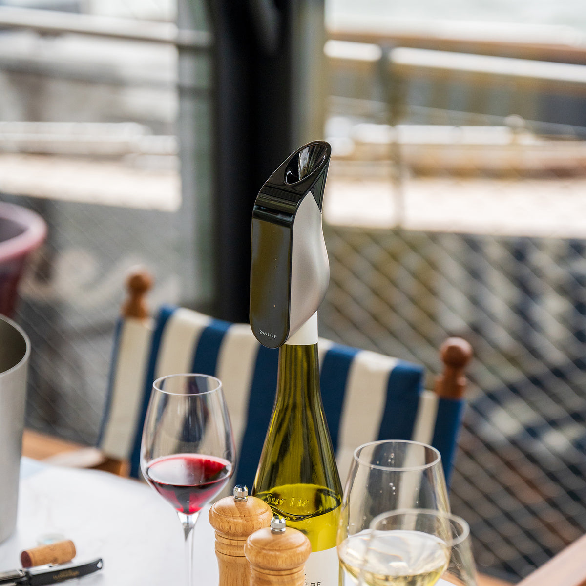 A bottle of wine with the Aveine Original Smart Wine Aerator – Instant 1–24 Hour Aeration by Aveine, in sleek black and silver, is attached to the bottle and sits on a table beside two wine glasses, pepper mills, and a corkscrew. A striped chair and a softly blurred outdoor background emphasize the effortless wine aeration experience delivered by Aveine’s innovative micro-oxygenation technology.
