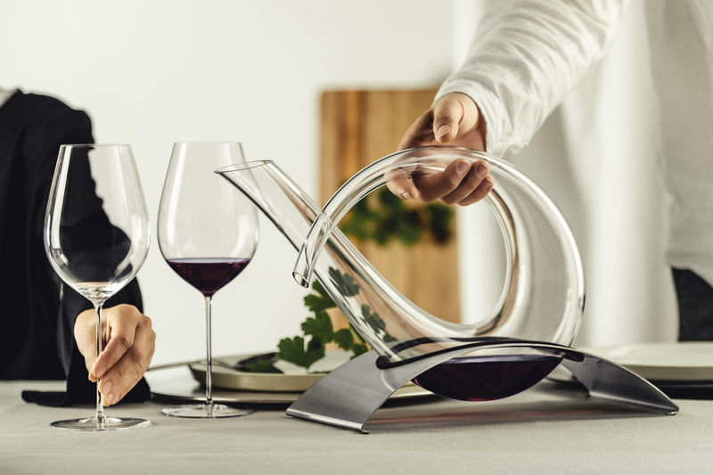 A modern wine decanter placed in a holder, sat on a table next to two red wine glasses.
