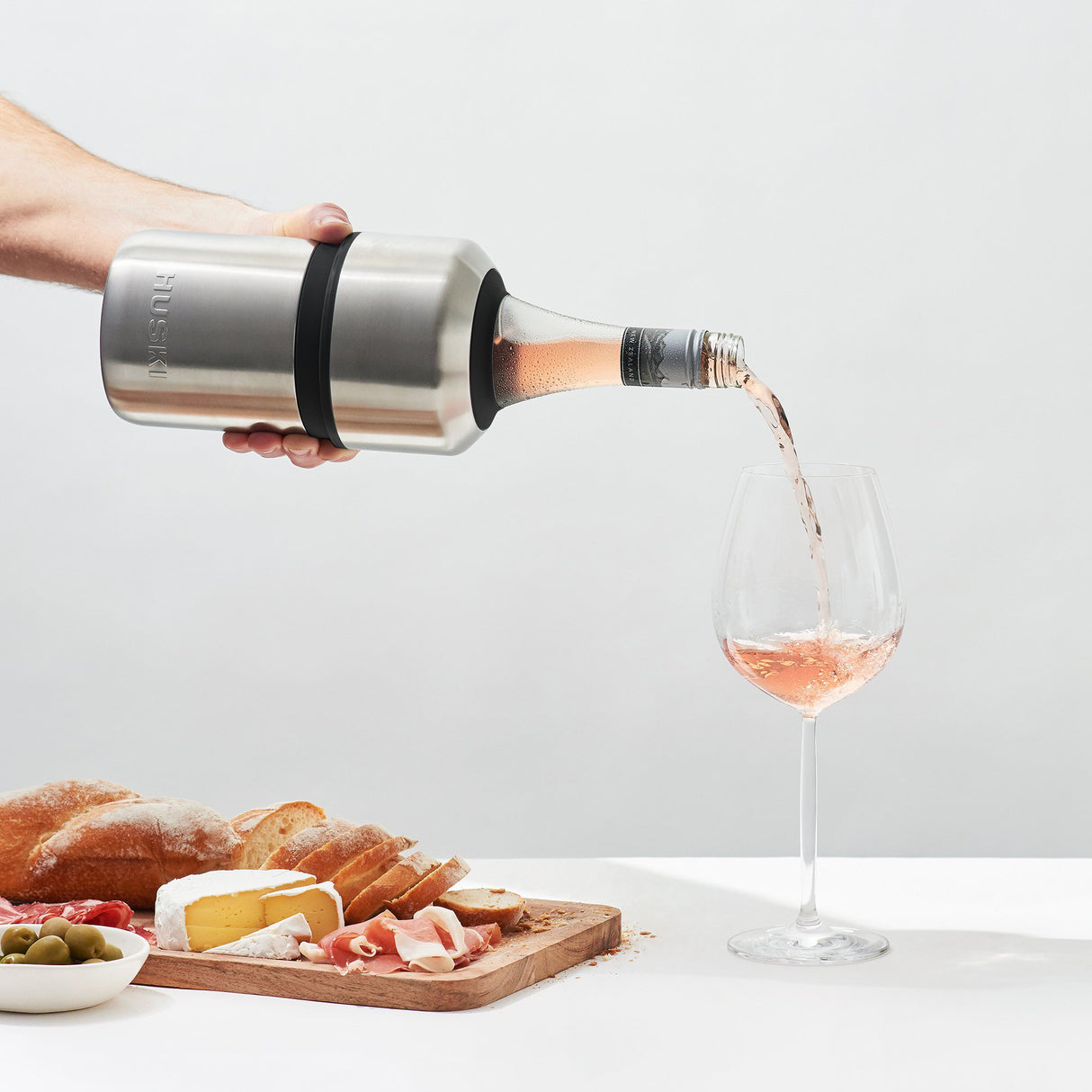 A hand pours rosé wine from a bottle kept chilled in the Huski Wine & Champagne Bottle Cooler by Huski, finished in brushed stainless steel, into a glass. Nearby, a wooden board with bread, cheese, meats, and olives rests against a plain white background.
