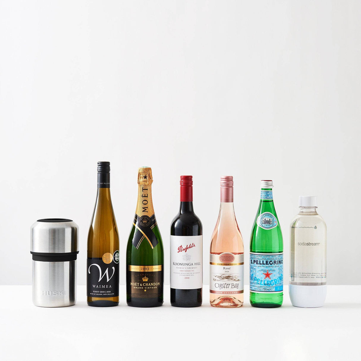 Six bottles—including wine, champagne, rosé, sparkling water, and a soda maker bottle—are lined up in a row with the Huski Wine & Champagne Bottle Cooler by Huski in brushed stainless steel positioned on the left, all set against a white background.