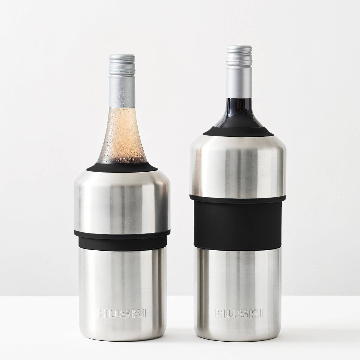 Two Huski Wine & Champagne Bottle Coolers by Huski, crafted in brushed stainless steel with black grips, each securely holding a wine bottle topped with a silver cap, are displayed on a white surface against a plain light background—an elegant solution for keeping your wine or champagne perfectly chilled for any occasion.