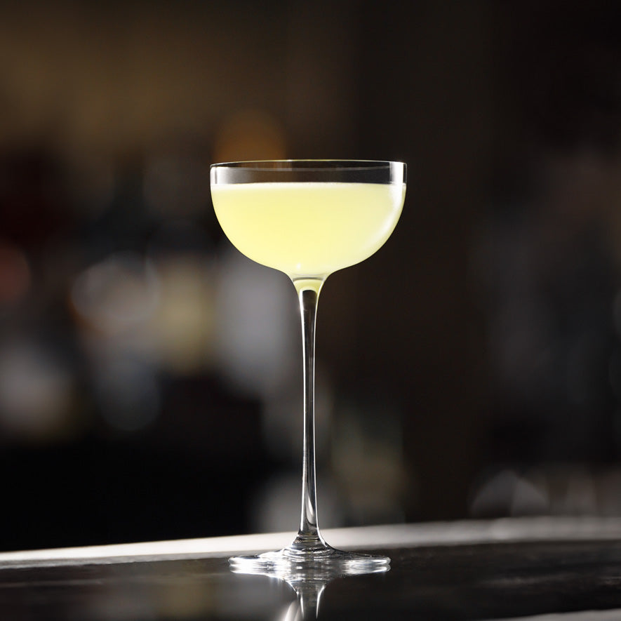 A coupe glass from the Lehmann Glass La Creative London Cocktail Coupe 200ml set evokes elegance as it cradles a pale yellow cocktail, resting on a polished bar surface against a softly blurred background.