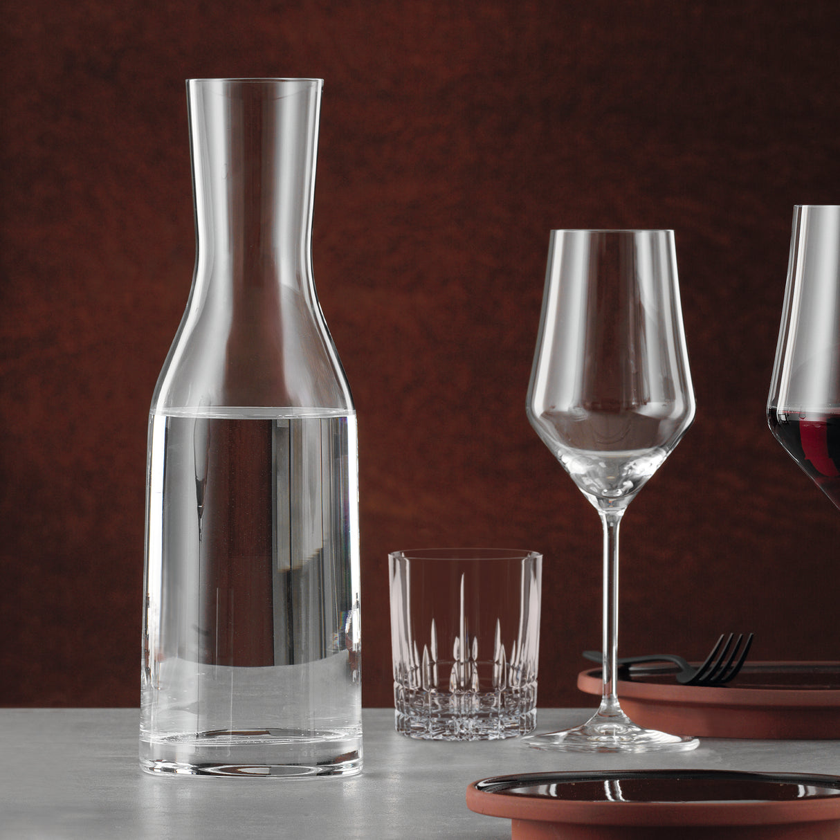 A Spiegelau Ultra Wine / Water Carafe 1000ml, filled with water, sits beside an empty tumbler, a wine glass, and a partially visible glass of red wine on a table set with plates and cutlery against a brown background. The elegant setup showcases the crystal clarity of the Spiegelau Ultra Carafe by Spiegelau as the centerpiece.