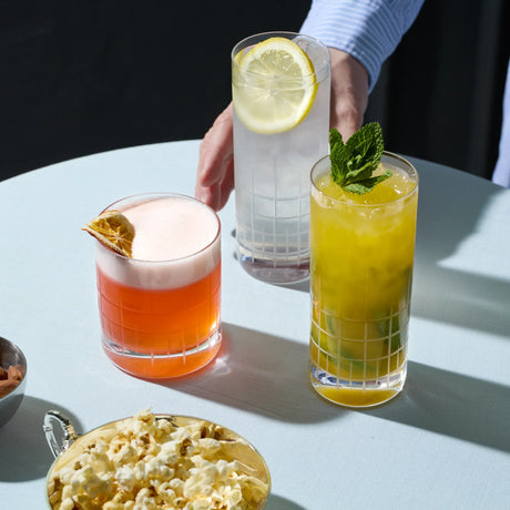 Three cocktails on a table with a bowl of popcorn and a hand reaching toward one of the glasses.