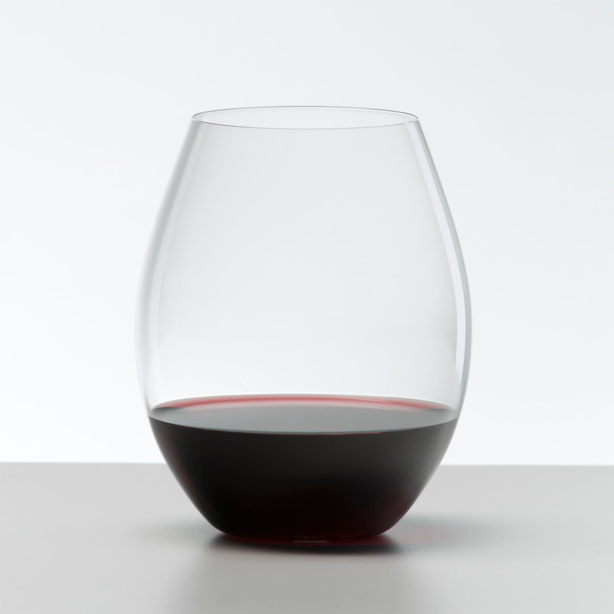 A Riedel O Range Stemless Old World Syrah Glass from Riedel, partially filled with red wine, rests on a light gray surface against a plain white background. This stylish set of two crystal glasses (0414/41) is both elegant and dishwasher safe.