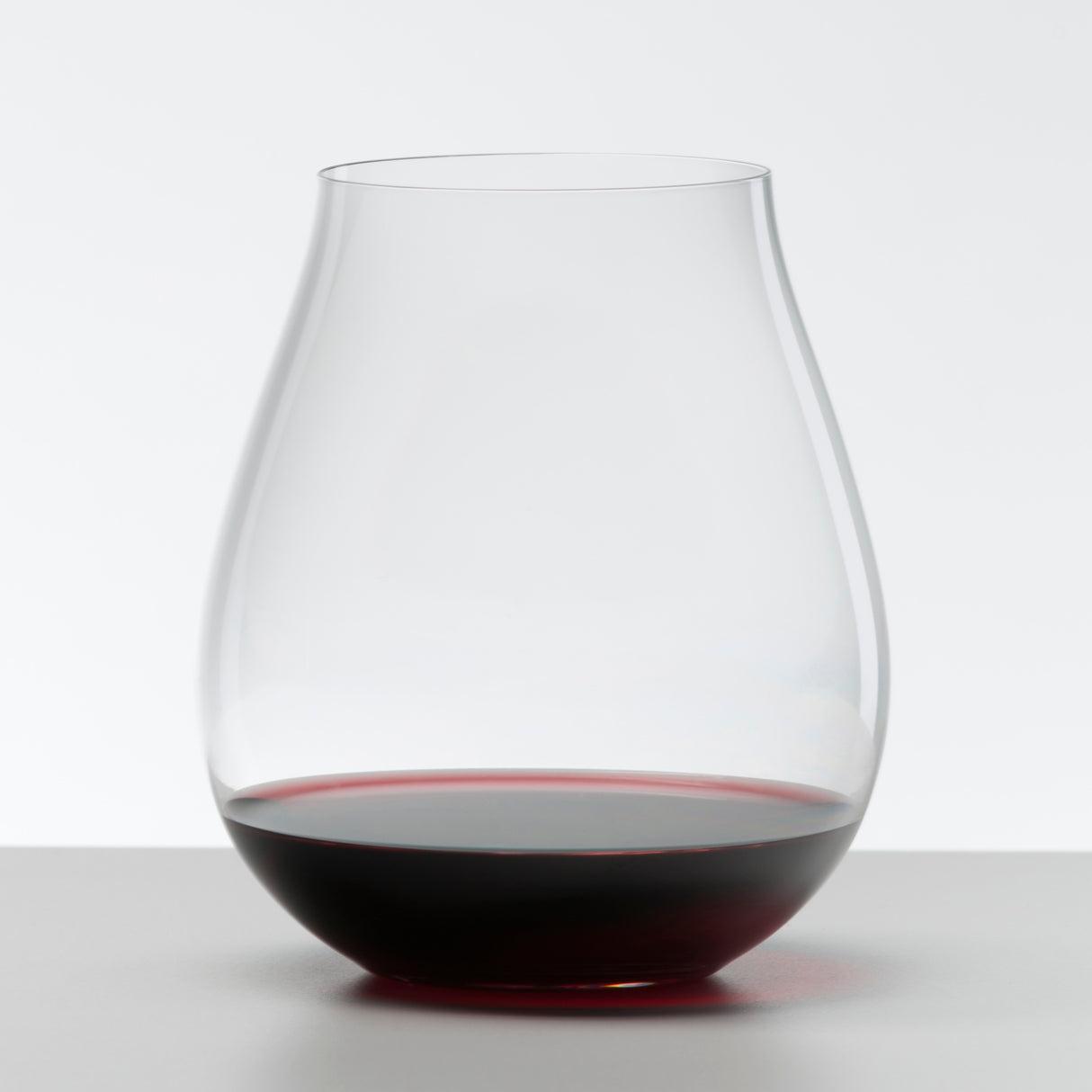 A clear, tulip-shaped stemless glass from the Riedel O Range Stemless New World Pinot Noir set (0414/67) by Riedel holds a small amount of red wine on a white surface with a plain background.