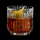 A Riedel Restaurant Bar Rituals Rocks Tumbler (283ml) from Riedel, filled with an amber-colored cocktail and ice cubes, garnished with a twisted lemon peel, set against a black background.