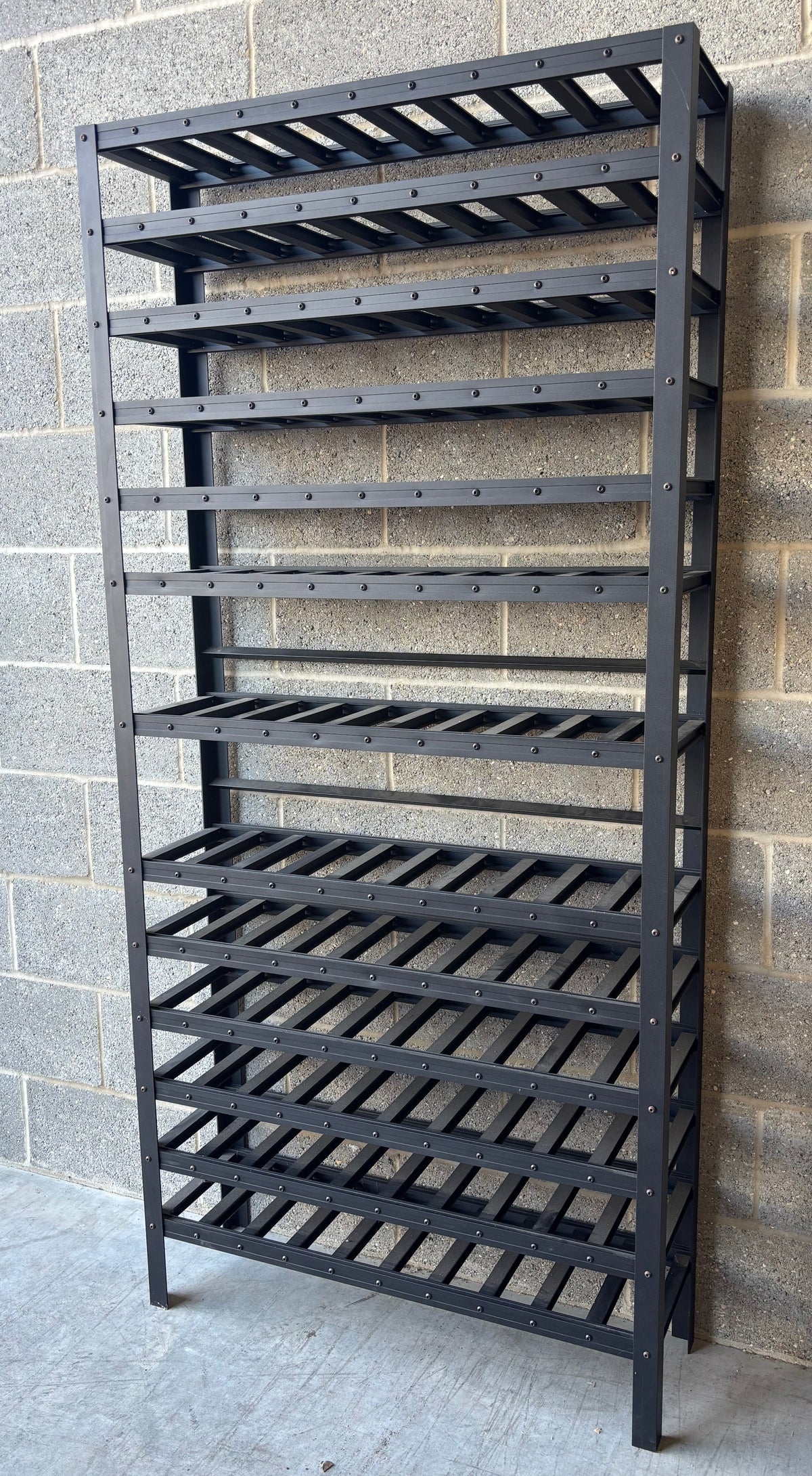 A tall black metal shelving unit with multiple slatted wine or bottle racks, standing against a gray cinderblock wall on a concrete floor.
