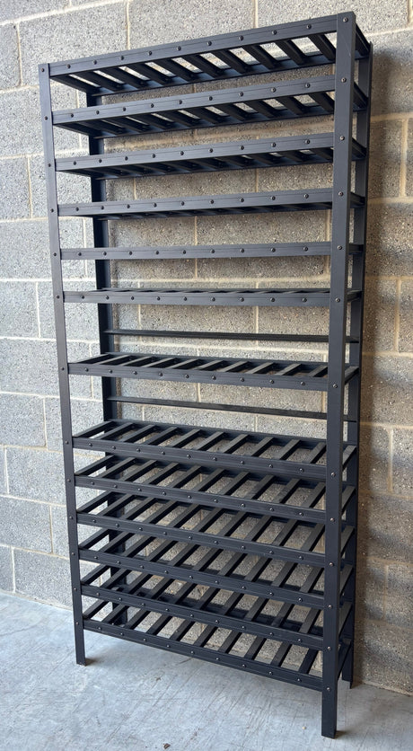 A tall black metal shelving unit with multiple slatted wine or bottle racks, standing against a gray cinderblock wall on a concrete floor.