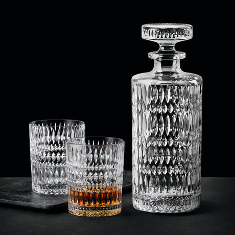 A Nachtmann Ethno Whisky Decanter & Two Glass Set by Nachtmann, featuring a clear, patterned decanter with a stopper and two matching crystal tumblers—one filled with amber whisky—are arranged against a dark background.