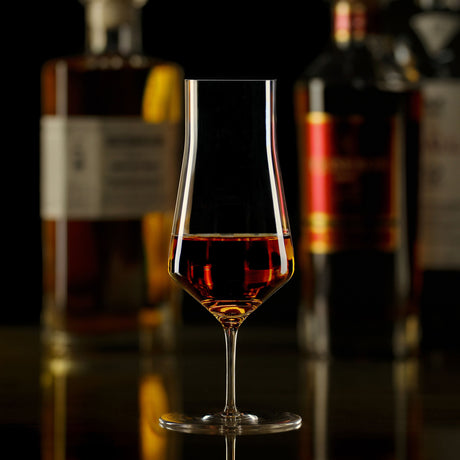 A Zalto Denk Art Cask Stemmed Spirit Glass filled with amber-colored liquid, positioned in front of two blurred liquor bottles.