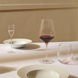 A table setting featuring a tall, angular Stolzle Quatrophil Red Wine Glass filled with wine, accompanied by a small stemmed glass, a short tumbler, and two wide-rimmed bowls.
