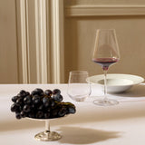 A stemless glass, a tall Stölzle Quatrophil Bordeaux Red Wine Glass filled with red wine, and a cluster of black grapes in a silver dish on a white tablecloth.