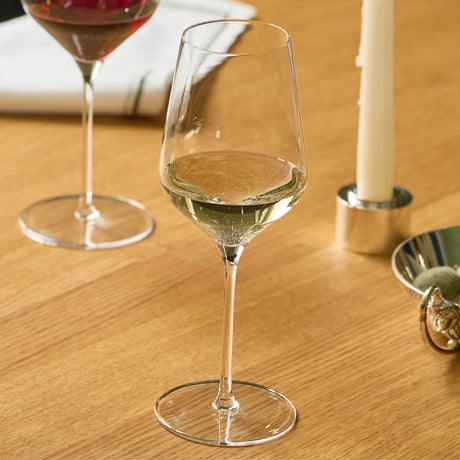 A clear stemmed Stolzle STARlight White Wine Glass partially filled with wine, standing on a wooden table next to a taller wine glass, a candle, and a small dish.