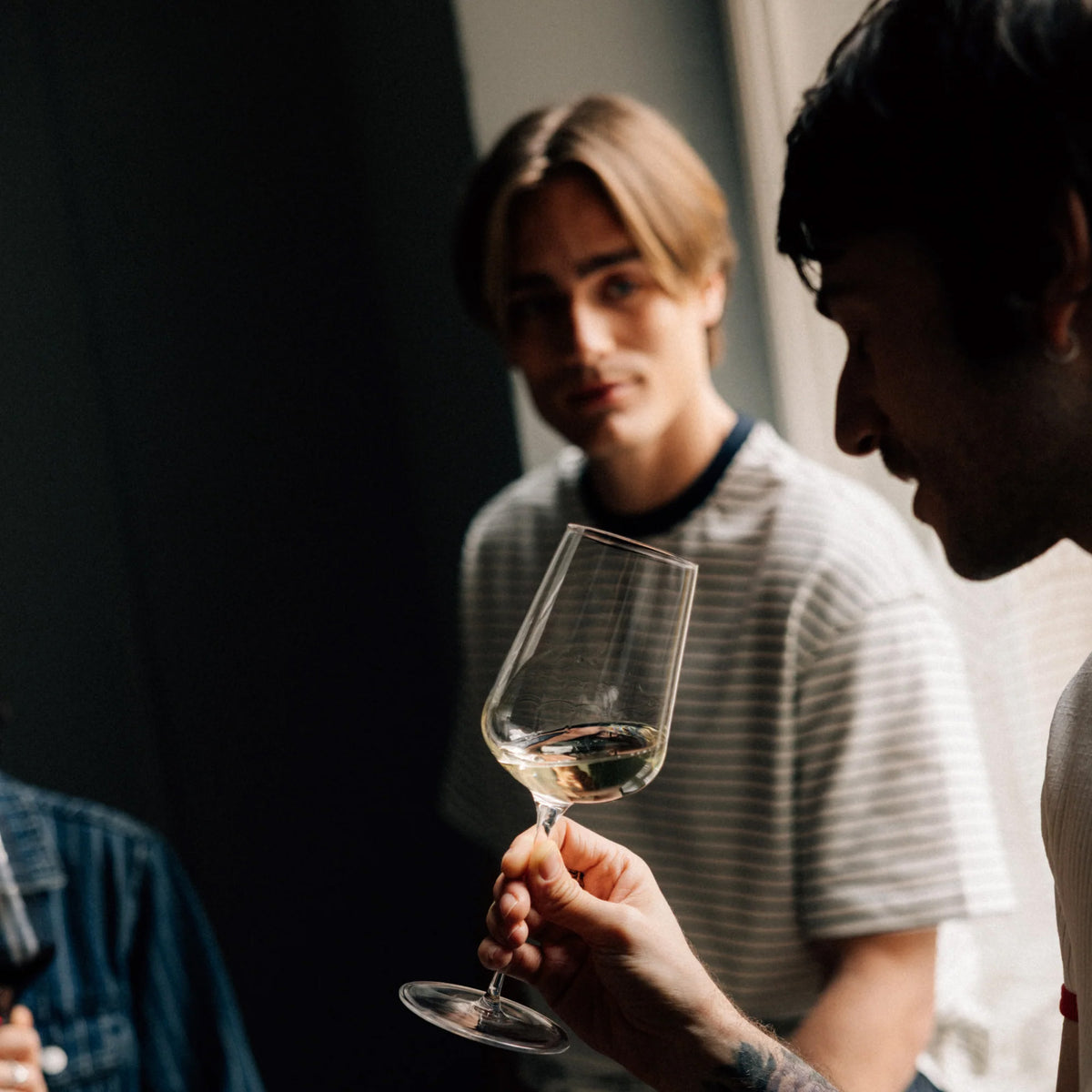 A person holds a clear, dishwasher-safe Stölzle STARlight Universal Red & White Wine Glass filled with white wine, with two people looking on in the background.