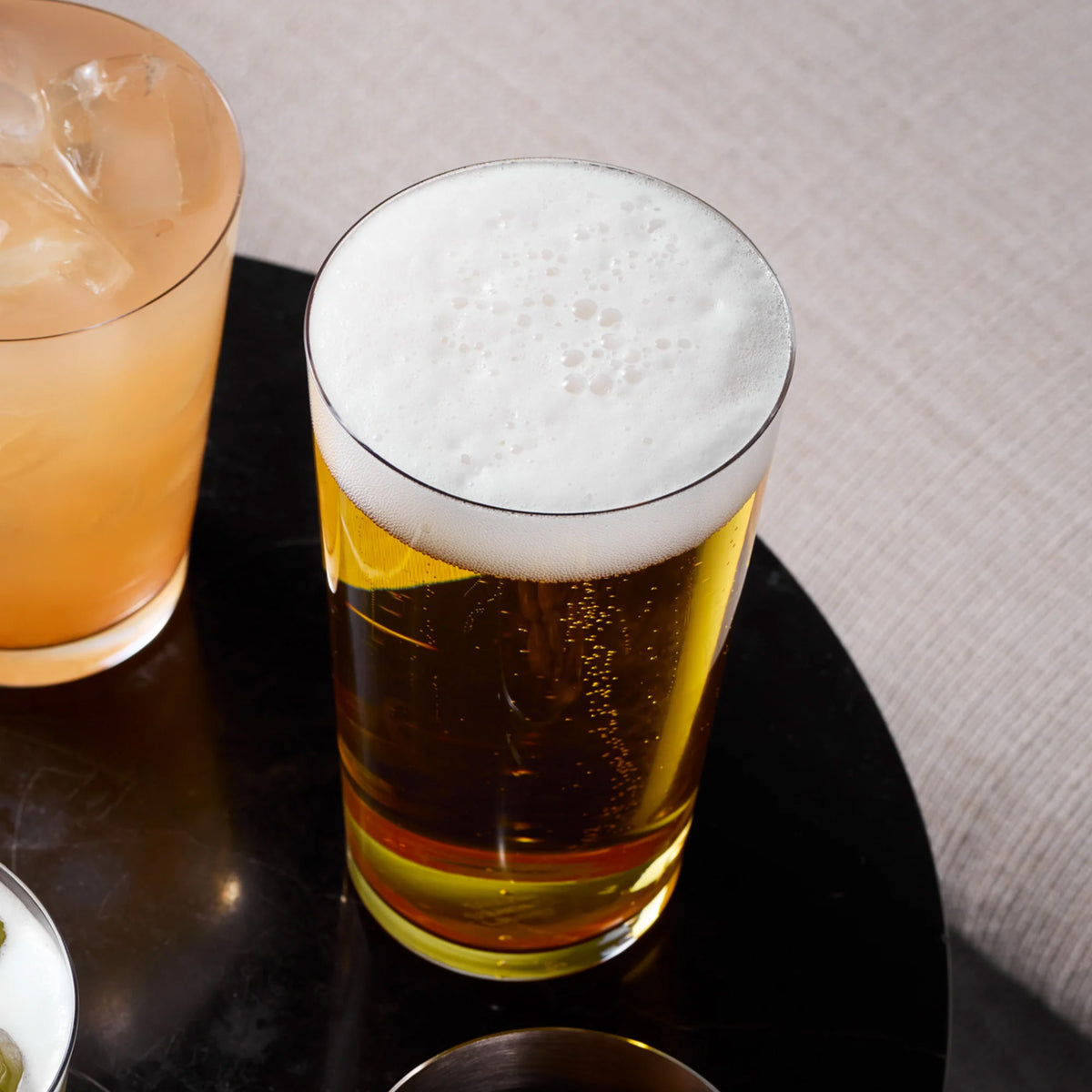 A tall, straight-sided Stölzle Metrico Beer Glass / Large Tumbler filled with amber beer and topped with a thick head of foam, sitting on a dark surface next to a glass of iced beverage.