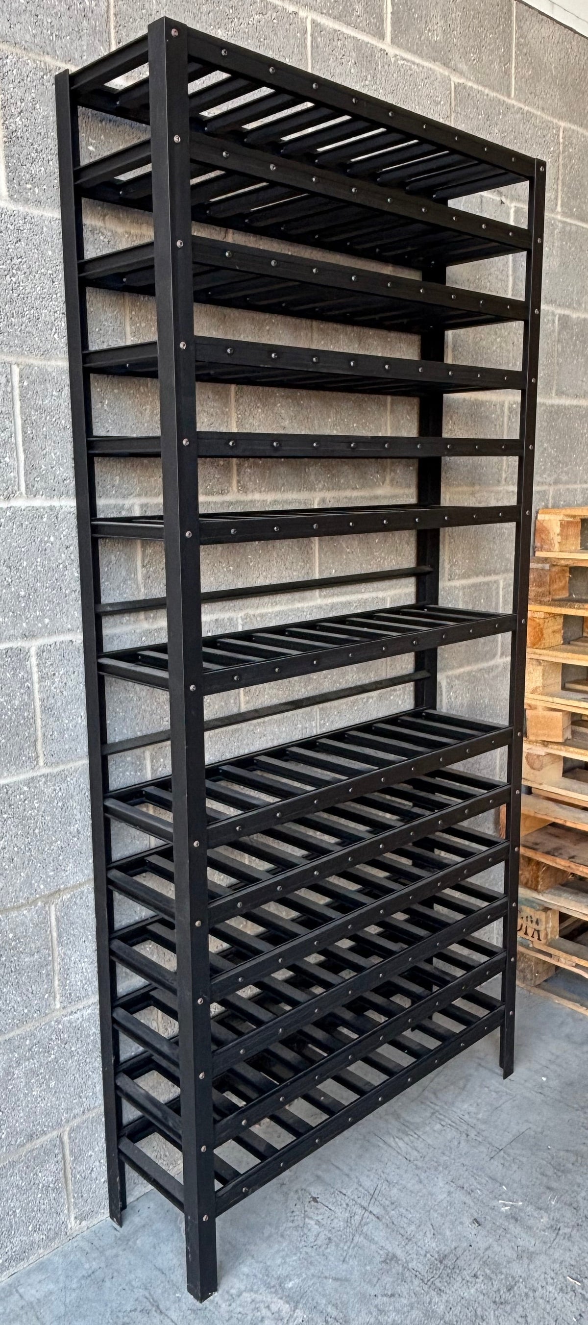 A tall black metal wine rack with multiple slatted shelves, photographed in a warehouse-like setting against a gray cinderblock wall with wooden pallets visible on the right.