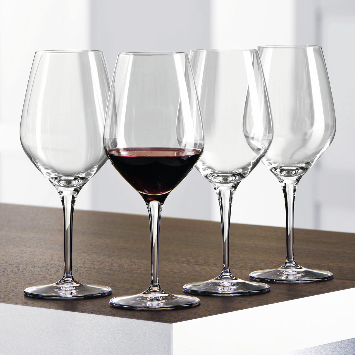 Twelve Spiegelau Restaurant Authentis Red Wine Glasses (480ml) by Spiegelau are arranged on a wooden surface; three glasses are empty while one is filled with red wine. The softly blurred white and gray background accentuates the professional design of these premium wine glasses.