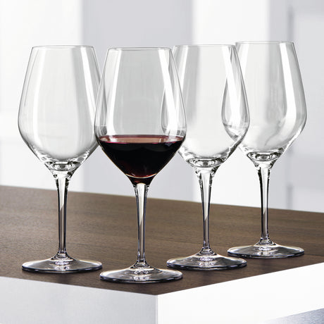 Twelve Spiegelau Restaurant Authentis Red Wine Glasses (480ml) by Spiegelau are arranged on a wooden surface; three glasses are empty while one is filled with red wine. The softly blurred white and gray background accentuates the professional design of these premium wine glasses.