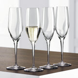 Twelve Spiegelau Restaurant Authentis Champagne Glasses (220ml) from Spiegelau are elegantly arranged on a wooden surface; three of the exquisite crystal glasses are empty and one is filled with sparkling champagne. The softly lit, out-of-focus background enhances the sophisticated presentation.
