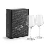 Two clear wine glasses—crafted from lead-free crystal by Gabriel-Glas—stand beside a sleek black rectangular box adorned with white script writing and a list of place names printed in small white text across the front. The Gabriel-Glas StandArt Universal Red & White Wine Glass Set of 2 offers an elegant, versatile design ideal for any occasion.