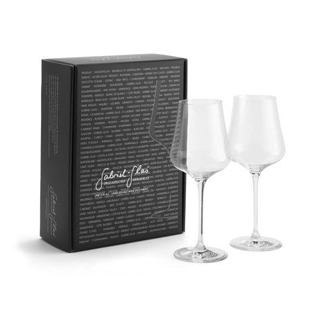 Two clear wine glasses—crafted from lead-free crystal by Gabriel-Glas—stand beside a sleek black rectangular box adorned with white script writing and a list of place names printed in small white text across the front. The Gabriel-Glas StandArt Universal Red & White Wine Glass Set of 2 offers an elegant, versatile design ideal for any occasion.