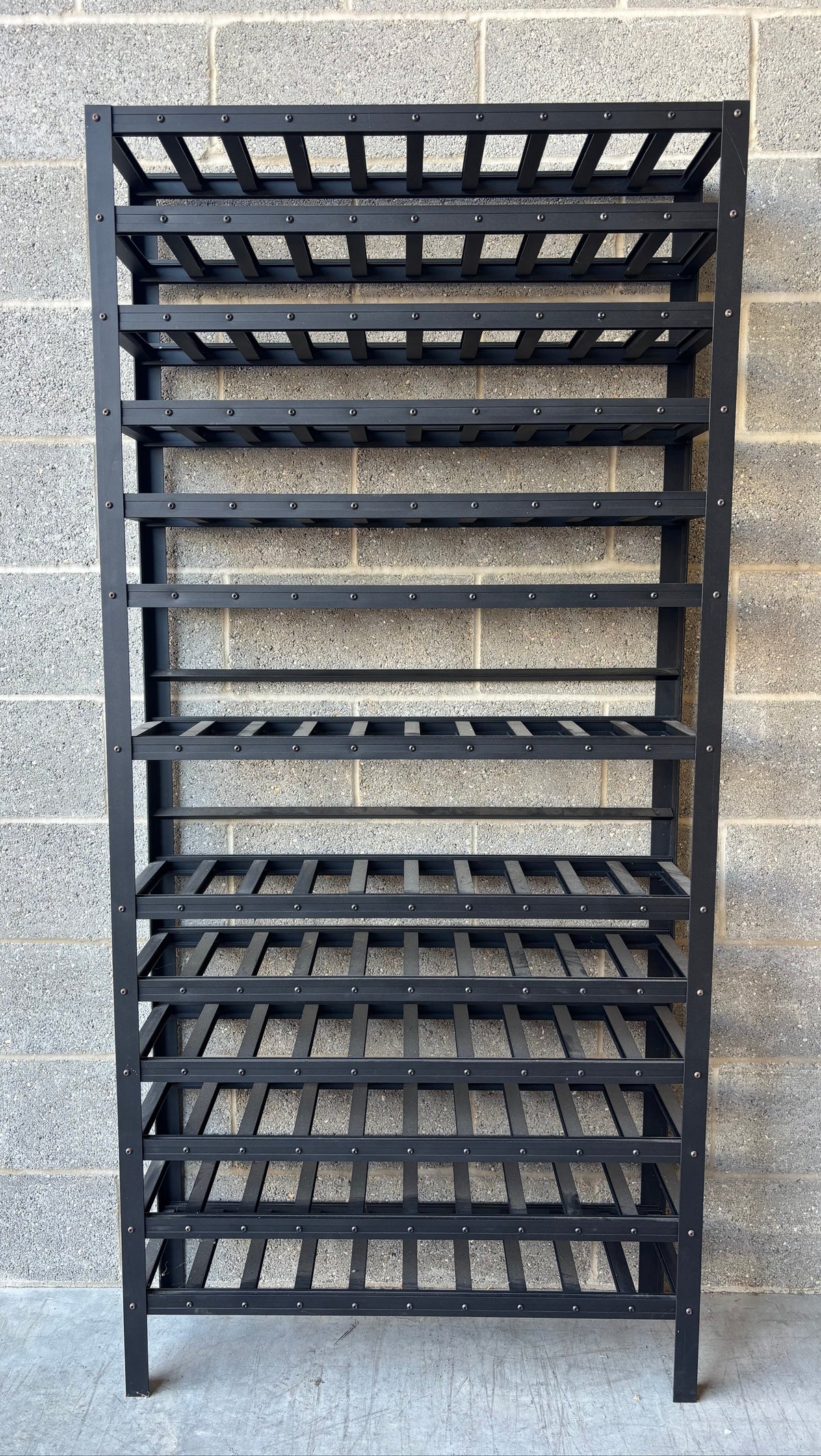 A black metal wine rack with multiple slatted shelves, standing against a gray concrete block wall.
