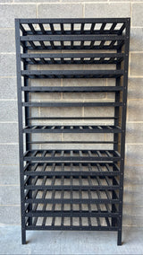 A black metal wine rack with multiple slatted shelves, standing against a gray concrete block wall.
