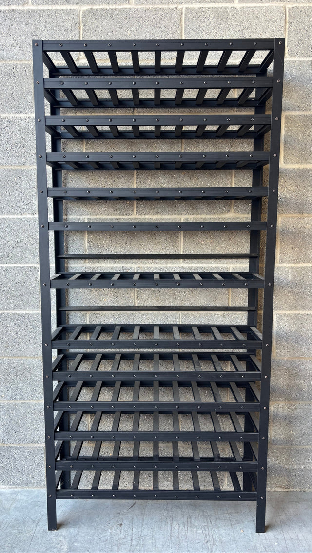 A black metal wine rack with multiple slatted shelves, standing against a gray concrete block wall.