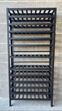 A black metal wine rack with multiple slatted shelves, standing against a gray concrete block wall.