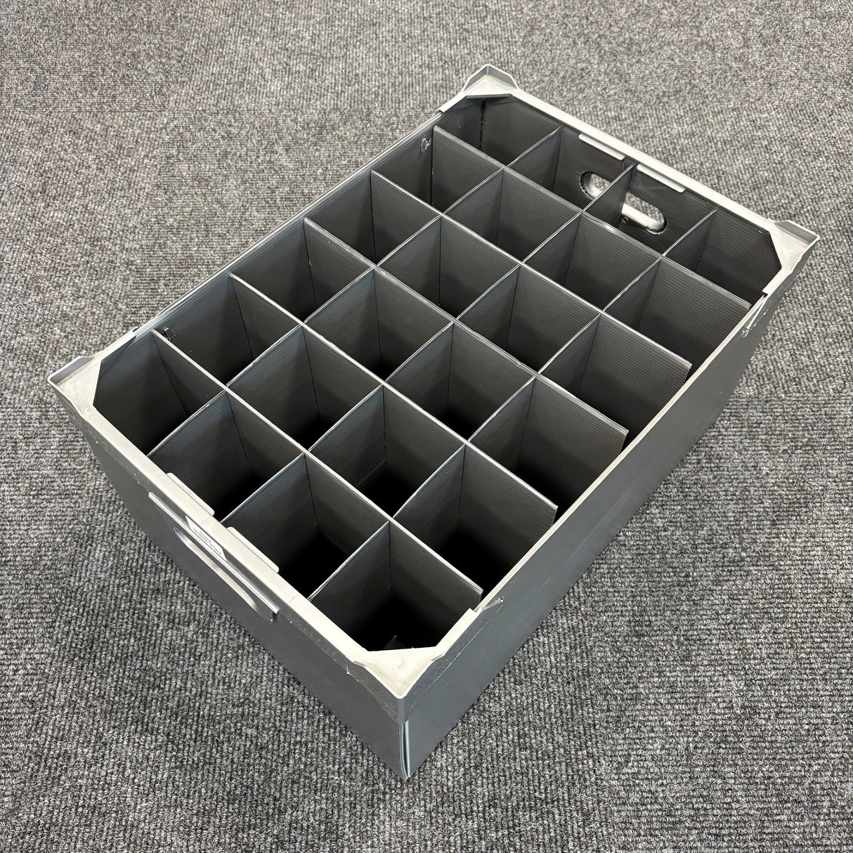 A USED - GREAT CONDITION Wine Glass Storage Box by Wineware, featuring 24 square compartments and measuring 230mm high, sits empty on a gray carpeted floor. The gray plastic box has handles on both ends and is ideal for securely storing wine glasses.
