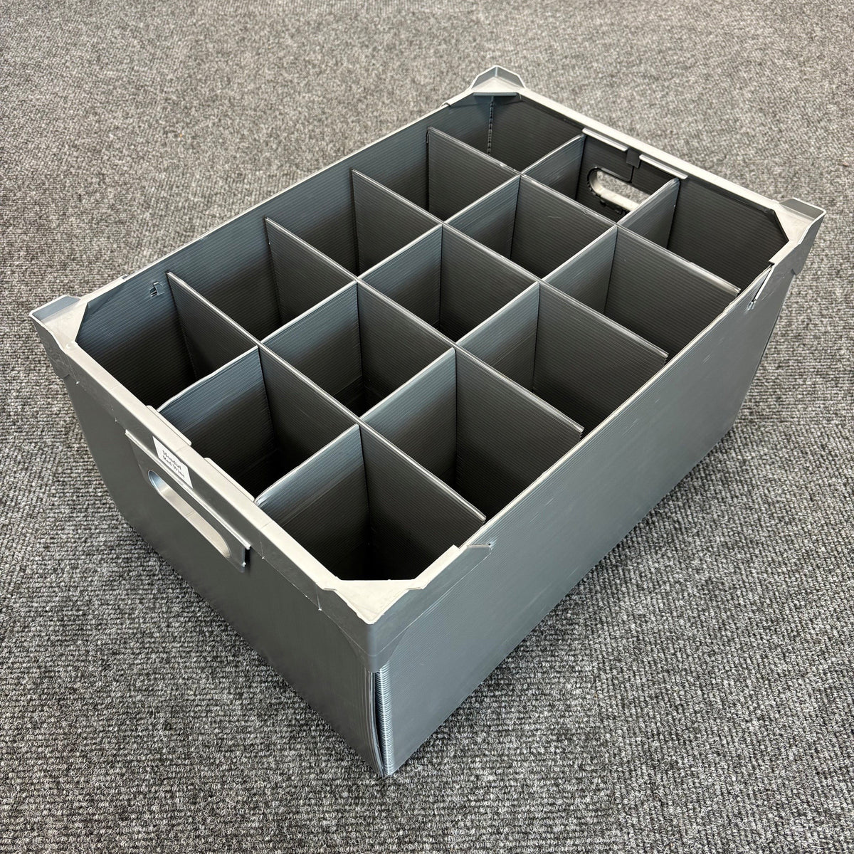 A Wineware USED - GREAT CONDITION Wine Glass Storage Box with 15 cells and measuring 240mm high is shown placed on a gray carpeted floor. Made of gray plastic, the box features handles and rectangular dividers that create a grid inside, making it ideal for use as a durable glassware or wine glass storage box.